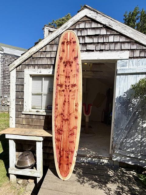 A finished wooden surfboard standing at the door of a shingled shack
