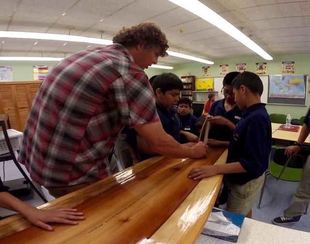 Rich showing a wooden surfboard to students who are touching the deck