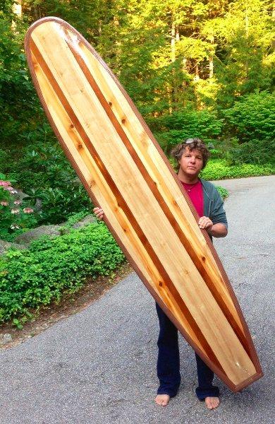 A young person standing beside a finished wooden surfboard taller than they are