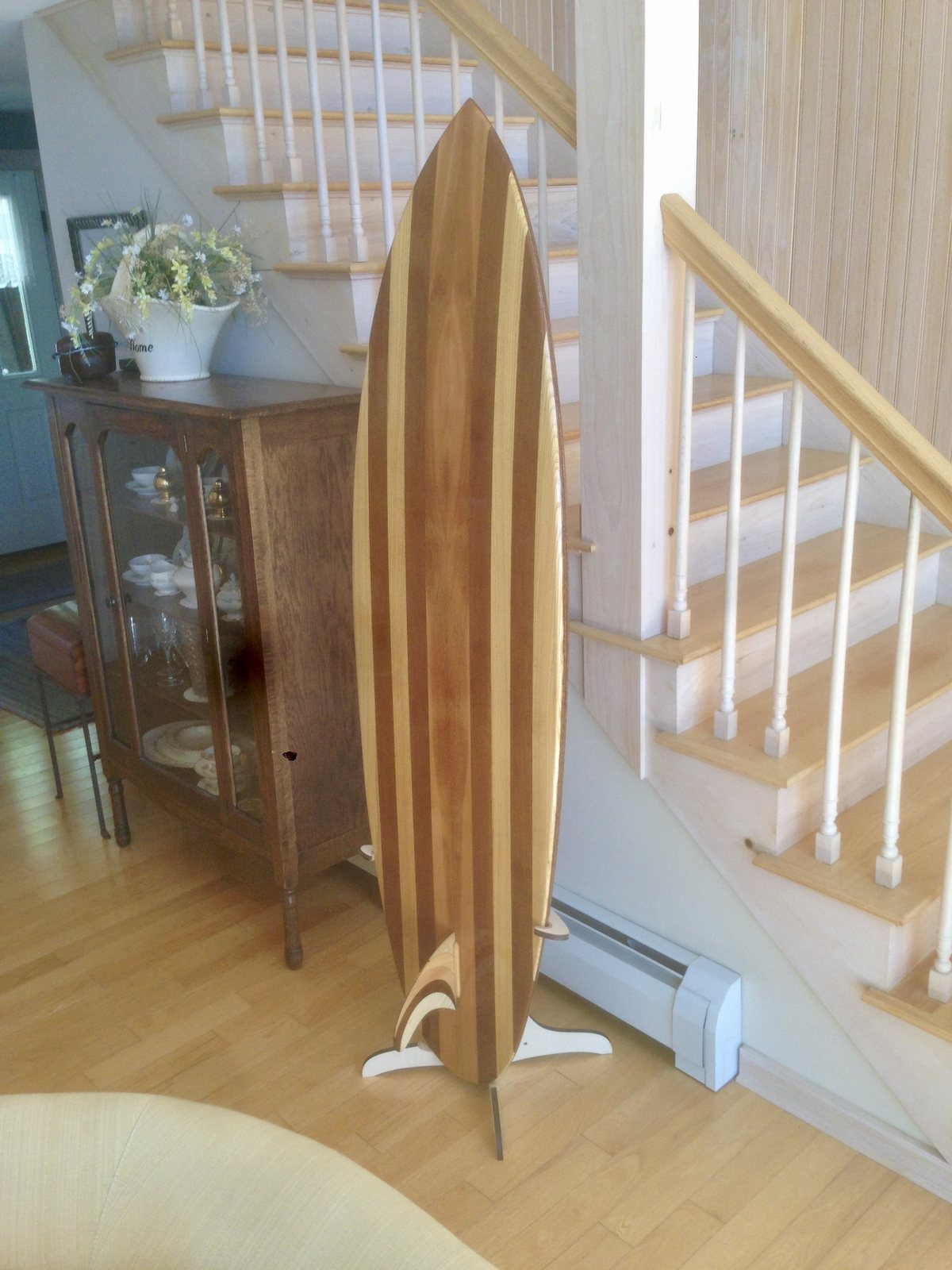 A striped wooden surfboard on a free-standing display stand at the foot of a staircase, fin set into the base