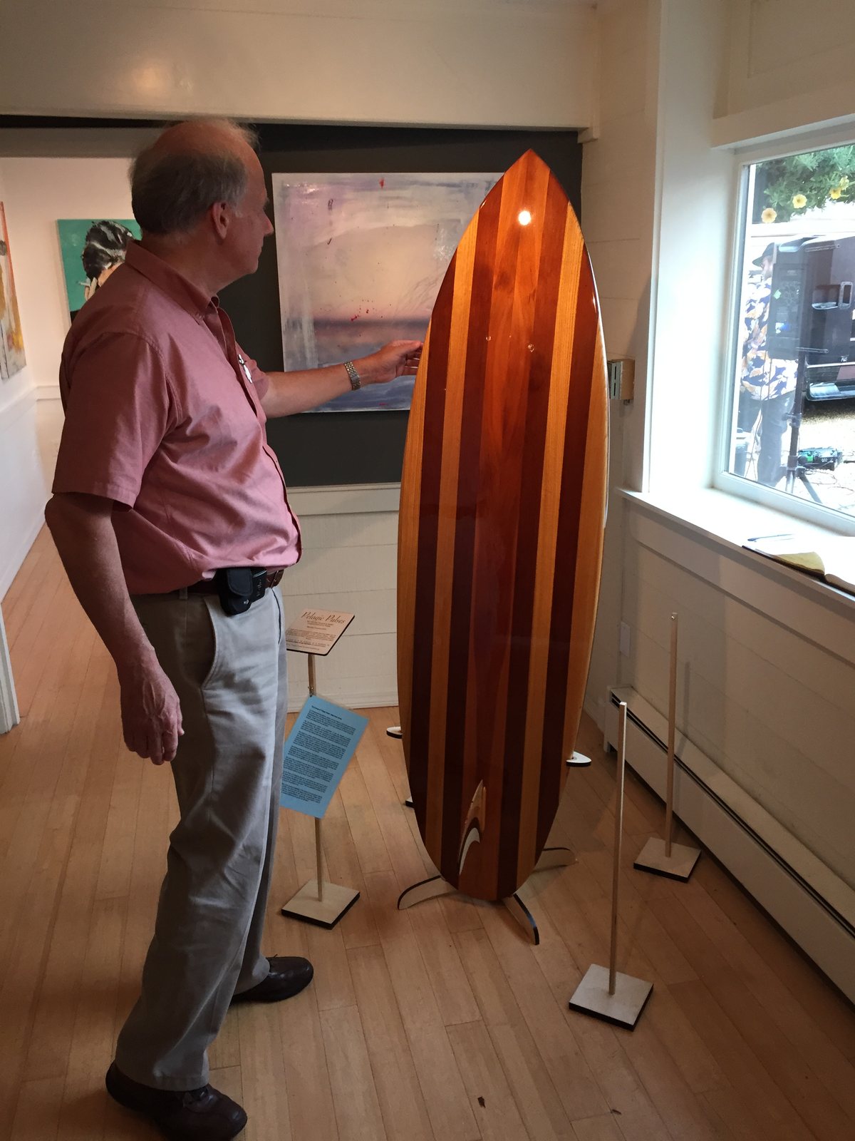 A visitor studies a striped wooden surfboard standing on its display stand in a gallery