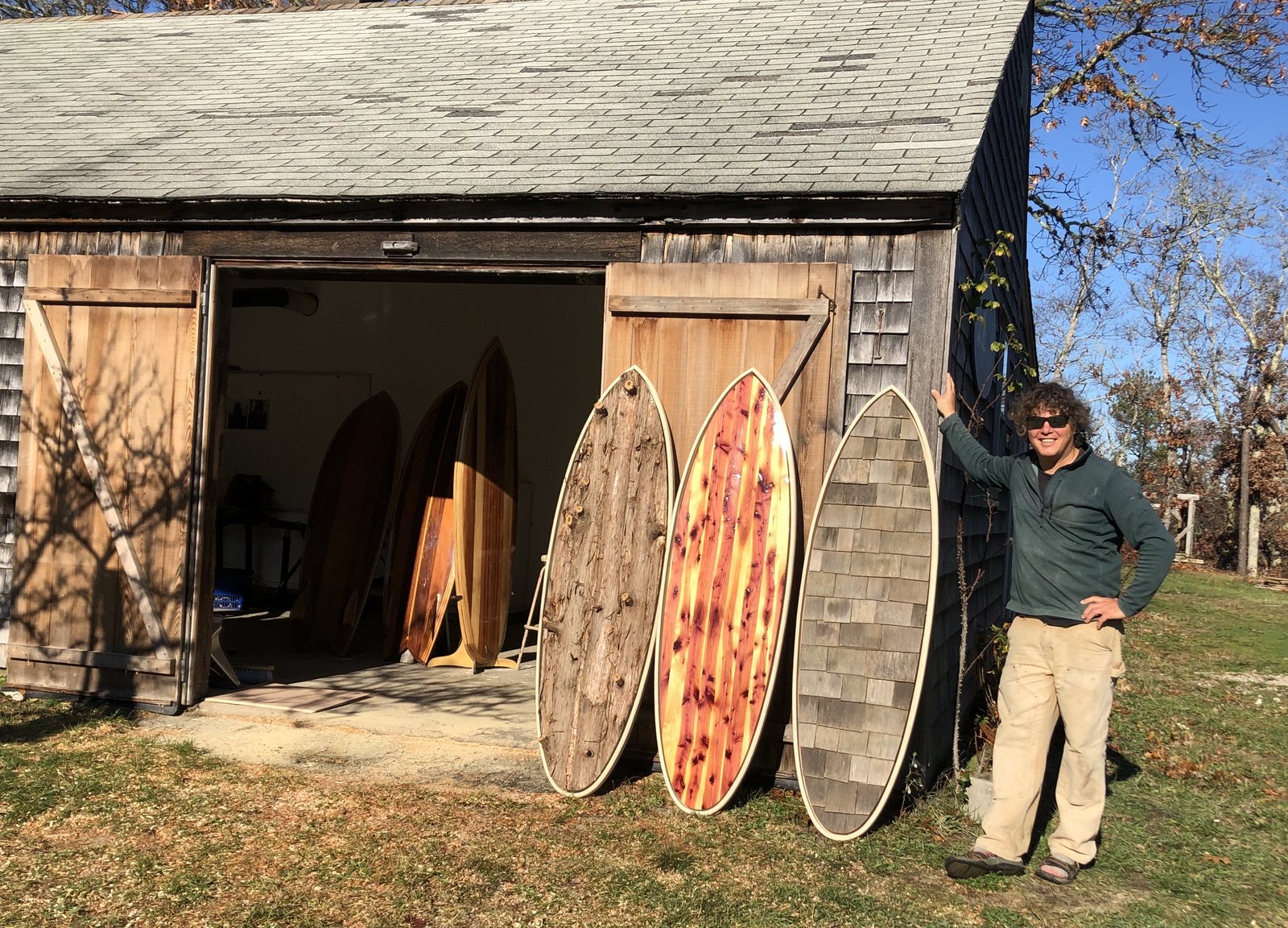 Rich beside three boards leaning at an open barn door: one faced in bark, one in cedar grain, one in shingles